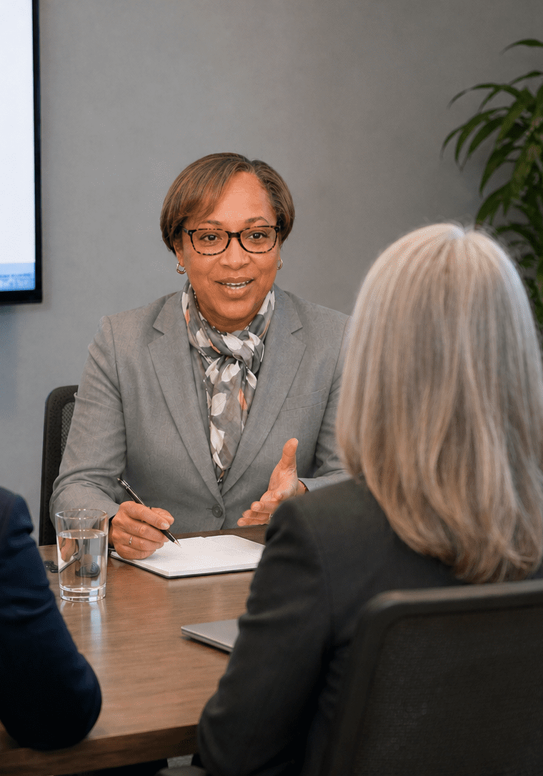 Business leader facilitating a conference room discussion with three colleagues, with a presentation screen about AI technology for language assessment and feedback in the background.